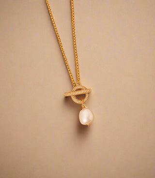 gold plated Cubic Zirconia encrusted T Bar & freshwater Pearl Necklace - Gold Lunar displayed on a concrete tile next to a crescent moon shaped pendant necklace and hammered band rings. sunlight shining on the jewellery.