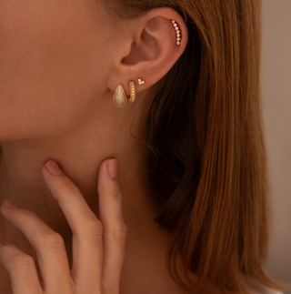 A person wearing gold huggie earrings adorned with cubic zirconia stones.
