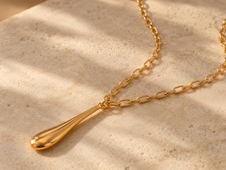 A gold teardrop pendant necklace with a matching chain, displayed on a textured beige stone surface with soft, dappled sunlight and shadows.