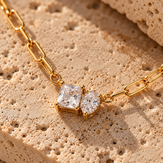 Gold chain necklace with a double-stone pendant featuring a square clear gemstone and a pear-shaped clear gemstone, displayed on a textured beige stone surface.