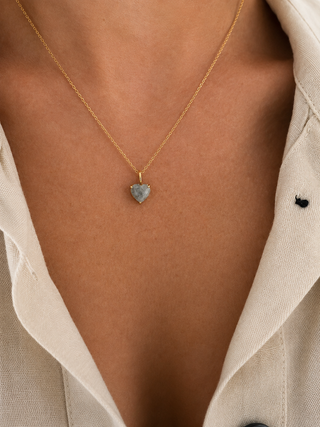 A close-up of a person wearing a delicate gold chain necklace with a small heart-shaped gray stone pendant, styled with a light beige linen shirt.