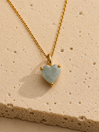 A gold heart-shaped pendant necklace with a pale greenish-blue stone, displayed on a textured beige stone surface.