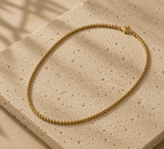 A gold beaded chain necklace with a heart-shaped clasp, displayed on a textured beige stone slab with soft leaf shadows.