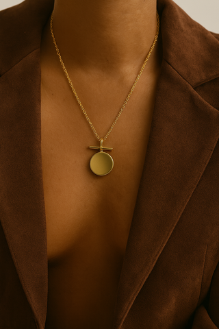 Gold necklace with a round pendant on a person wearing a brown jacket.