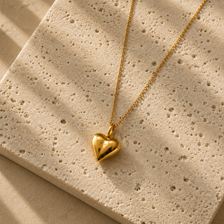 A polished gold heart pendant necklace on a delicate gold chain, displayed on a textured beige stone slab with soft diagonal shadows.