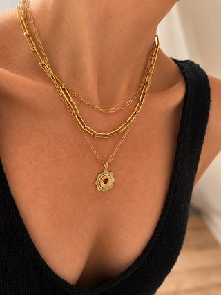 A close-up of a model wearing a layered gold necklace set, featuring a paperclip chain, a cable chain, and a delicate chain with a shield-shaped pendant containing a red heart in the center.