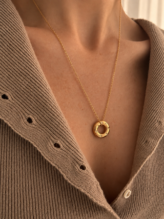 A close-up of a person wearing a delicate gold chain necklace with a textured, open ring pendant, styled over a brown knit cardigan.