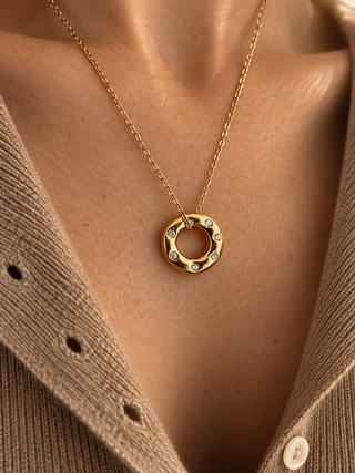 A gold-toned circular pendant necklace with small clear stones set along the lower half, worn on a model's neck over a beige knit cardigan.
