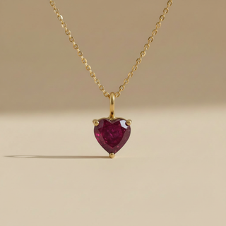 Gold necklace with a heart-shaped red gemstone pendant on a beige background