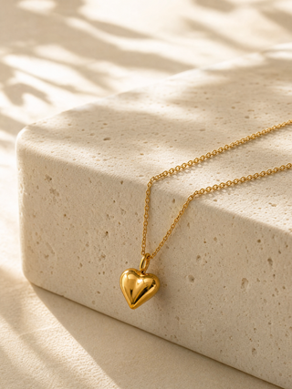 A gold heart pendant necklace on a delicate gold chain, draped over a textured beige stone block with soft, dappled sunlight and shadows.