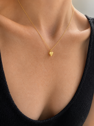 A close-up of a model's neck and chest wearing a delicate gold chain necklace with a small textured heart pendant, paired with a black knit top.
