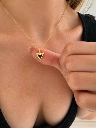 A close-up lifestyle shot of a model wearing a delicate gold chain necklace with a small heart-shaped pendant. The pendant features a polished gold border with a black center, held gently by the model's hand.