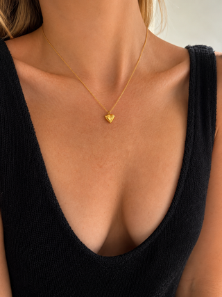 A close-up of a model wearing a delicate gold chain necklace with a small, polished heart-shaped pendant, styled with a black ribbed knit top.