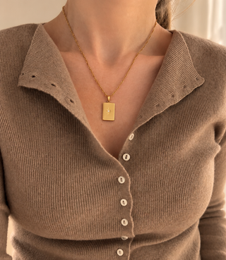 A close-up of a person wearing a taupe ribbed knit button-front cardigan with a pointed collar and a gold pendant necklace, photographed indoors near a window.