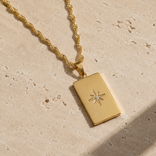 A gold rectangular pendant necklace with a starburst design and a small clear center stone, resting on a textured beige stone surface.