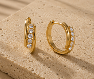 A pair of gold huggie hoop earrings with a row of clear pavé-set stones, displayed on a textured beige stone surface.