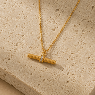 A gold necklace with a horizontal bar pendant featuring a small inset of clear stones, displayed on a textured beige stone surface.