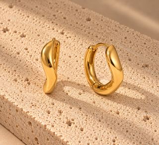 A pair of polished gold huggie hoop earrings with a unique organic wavy shape, displayed on a textured beige stone block.