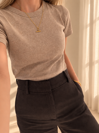 A person wearing a taupe heather short-sleeve crewneck t-shirt tucked into dark brown trousers, accessorized with a gold necklace and bracelet in a warm, sunlit indoor setting.