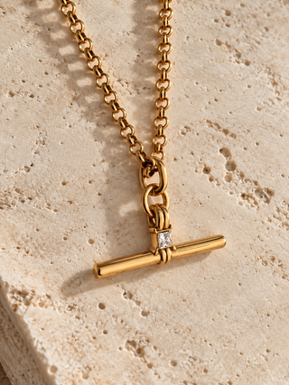 A gold chain necklace with a toggle bar pendant featuring a small clear gemstone, displayed on a textured beige stone surface.
