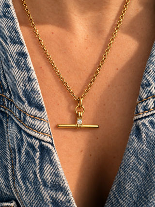 A gold chain necklace with a toggle bar pendant worn by a person in a denim jacket. The pendant features a small clear gemstone accent on the left side of the bar.