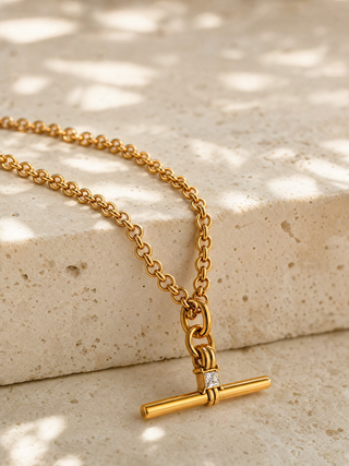 A gold chain necklace with a toggle bar pendant featuring a small clear stone, displayed on a textured beige stone surface with dappled sunlight.