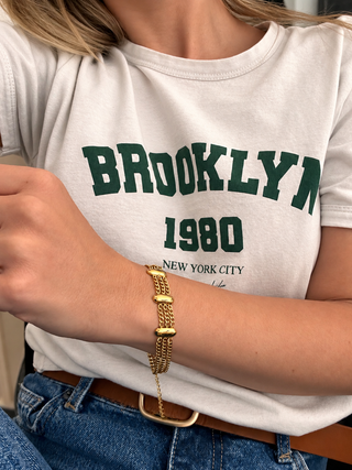 A close-up of a person wearing a white graphic T-shirt with green 'BROOKLYN 1980' text and a gold chain bracelet on the wrist, paired with blue jeans and a brown belt.