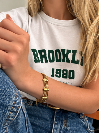 A close-up of a person wearing a white graphic T-shirt with green 'BROOKLYN 1980' text and a gold chain bracelet on the wrist, paired with blue jeans.