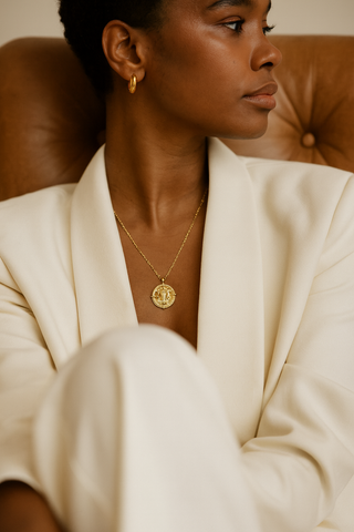 Zodiac gold necklace worn on a model sitting on a brown leather chair and wearing an ivory suit